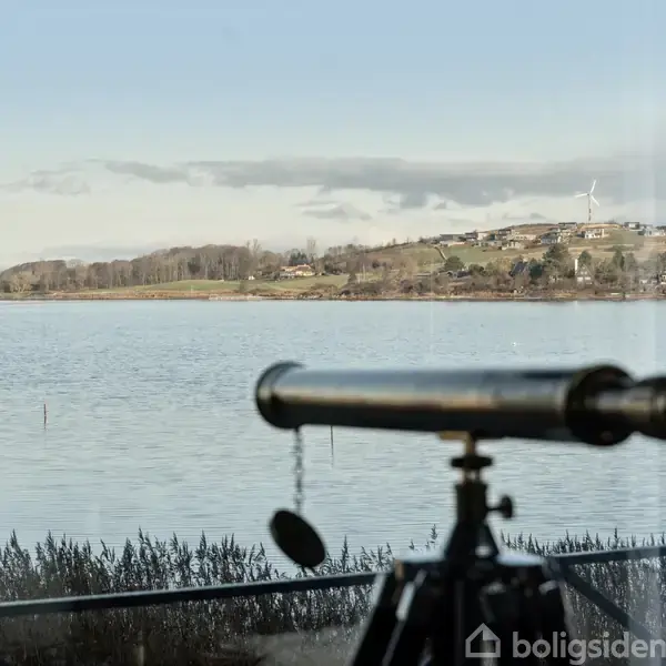 Et teleskop står foran et vindue og peger ud mod en sø. I baggrunden ses en bakkekam med et vindmølle og spredte bygninger.