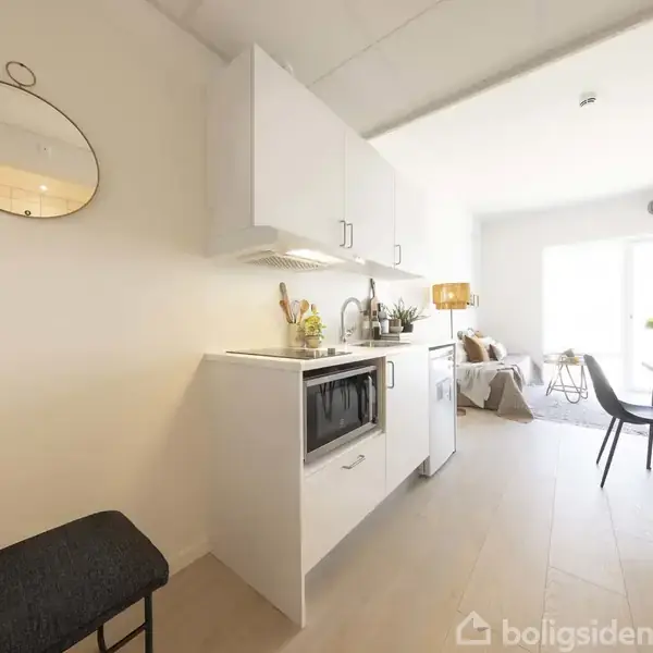 Et lille køkken med hvide skabe og en indbygget mikrobølgeovn. I baggrunden ses en stue med spisebord, stole og en sofa ved et stort vindue, der giver naturligt lys.