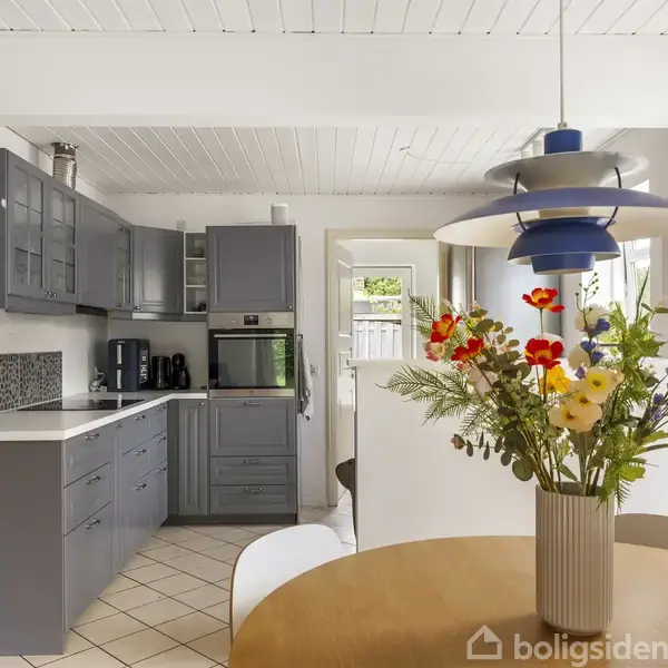 Køkken med grå skabe, ovn og kaffemaskiner. Rundt bord med vase af farverige blomster. Lampe hænger over bordet. Lyse vægge og flisegulv giver et lyst, moderne udtryk.