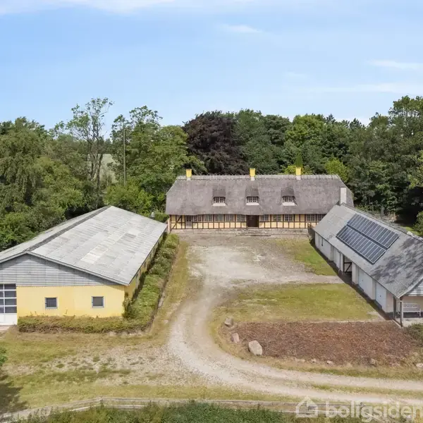Et bindingsværkshus med stråtag står i en solrig, grøn have. Huset har gule vægge, mange små vinduer og tre gule skorstene. Patiområdet omgiver huset med brostensbelægning.