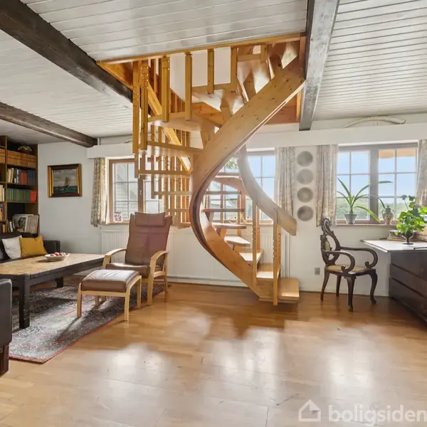 En trævindeltrappe står centralt i en stue med trægulv, omgivet af en sort lædersofa, reol fyldt med bøger og stort vindue med lyse gardiner, der slipper sollys ind.