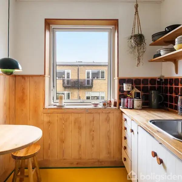 Et lille køkken med træpaneler har et rundt bord og stol ved vinduet. Vask og køkkentilbehør er til højre, med hylder ovenover. En lampe hænger over bordet.