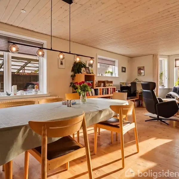 Et spisebord med stole står dækket med en dug, omgivet af et lyst opholdsrum med store vinduer, lamper hængende over bordet og en sofaarrangement i baggrunden.