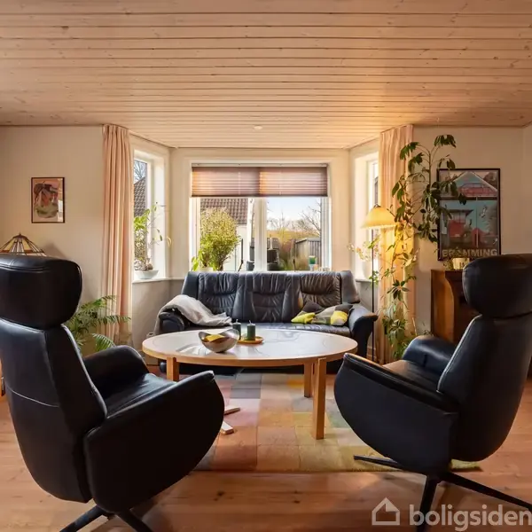 To sorte lænestole står foran et rundt sofabord. En sort sofa står ved det store vindue med udsigt til en have. Et TV til venstre og planter i hjørnet.