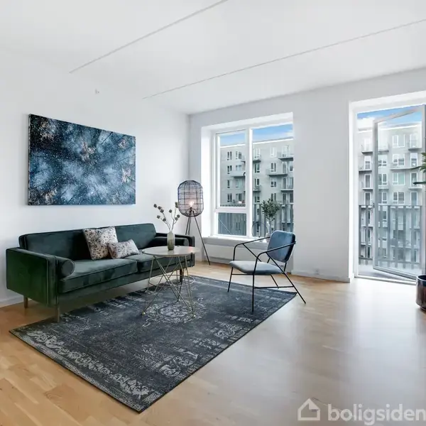 En grøn sofa står nær et stort vindue, flankeret af en moderne lampe og en stol. Indretningen suppleres med et tæppe, planter og vægkunst. Udsigt til bygningskomplekser udenfor.