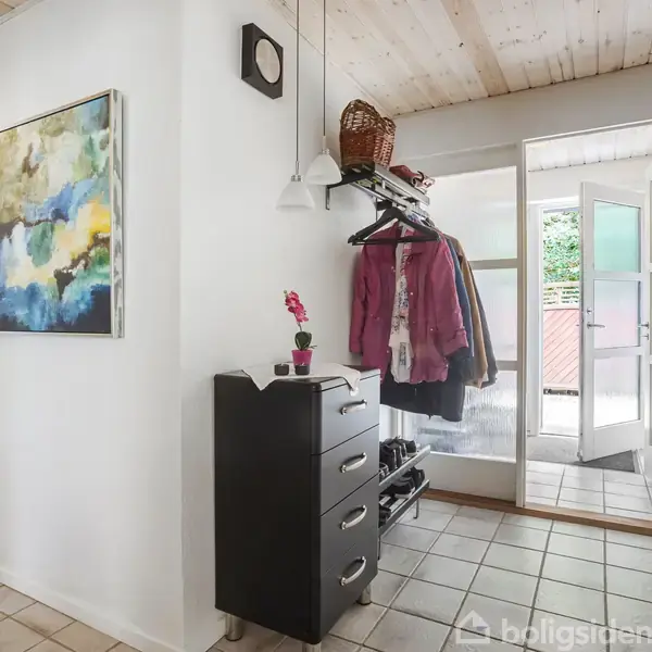 En entre med en kommode, hvor en lille plante står, jakker hænger på en knagerække med sko under, to malerier på væggen og åbne døre mod en lys veranda.
