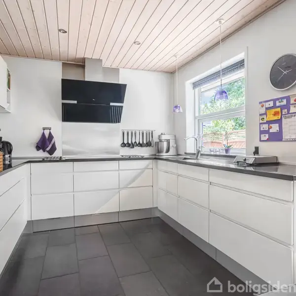 Hvidt, moderne køkken med indbyggede skabe og ovn; vægmonteret emhætte og kogeblus. På bordet står køkkenudstyr. Væggen har en opslagstavle og ur. Store vinduer slipper lys ind.