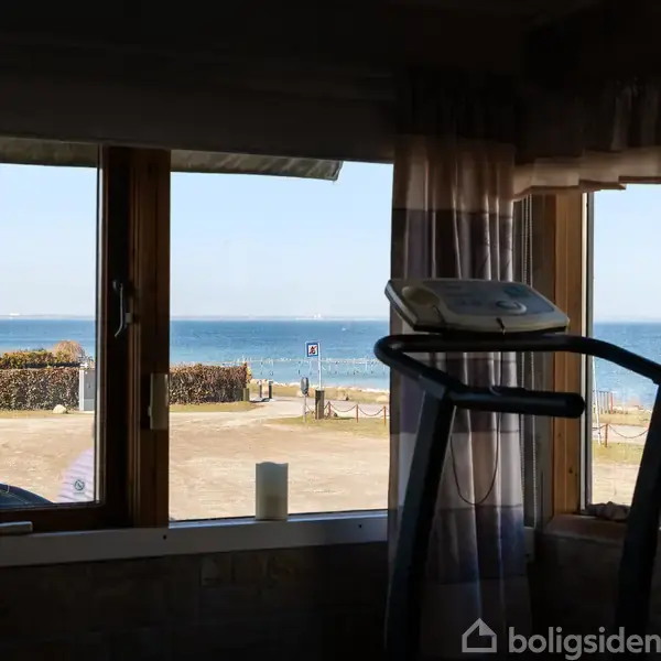 Løbebånd står stationært foran et stort vindue med udsigt til havet og en strand udenfor. Biler og skilte er synlige på parkeringspladsen.