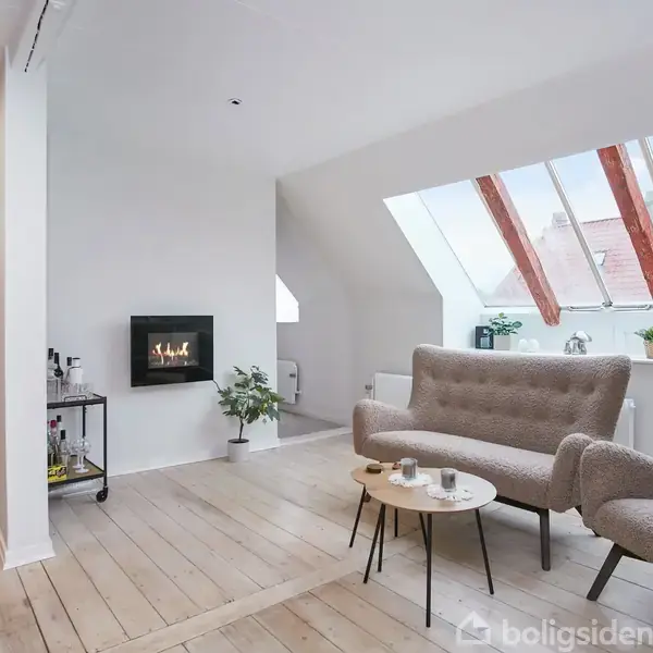 En stue med to polstrede stole og et småt rundt bord, oplyst af et stort tagvindue. En væghængt pejs brænder roligt. Trægulve og minimalistisk indretning dominerer rummet.