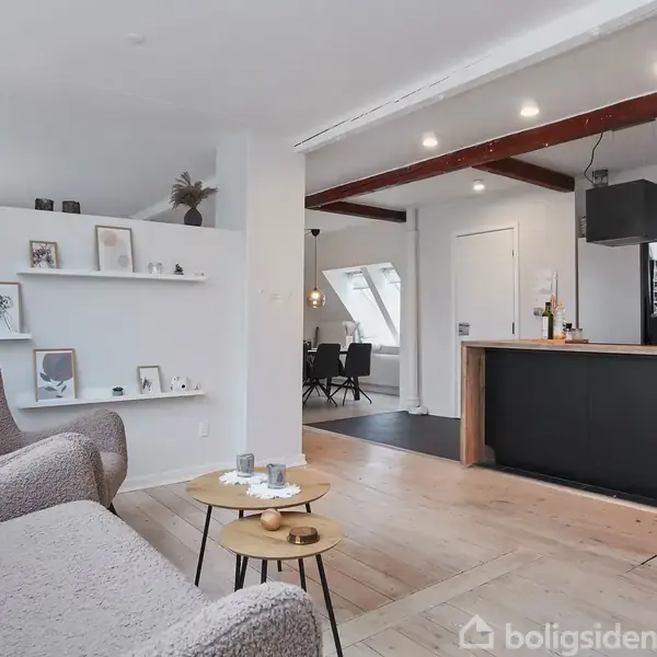 En hyggelig stue har to bløde lænestole, et lille rundt bord med te og et åbent køkken i moderne stil. Indretning inkluderer hylder med billeder.