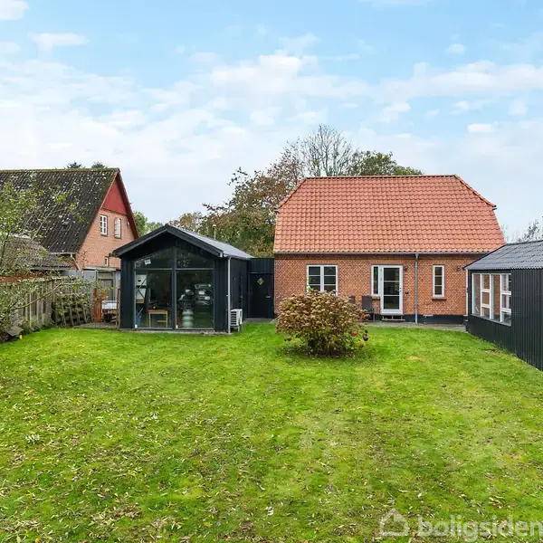 Hus står stille i en velholdt have med grøn græsplæne og træer. To bygninger ses: en rød murstenshus med orange tag og en sort træbygning til højre.