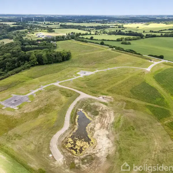 En grusvej snor sig gennem åbne marker med et lille vandhul i midten. Omkring ligger grønne marker og skove i et bakket landskab under en blå himmel.