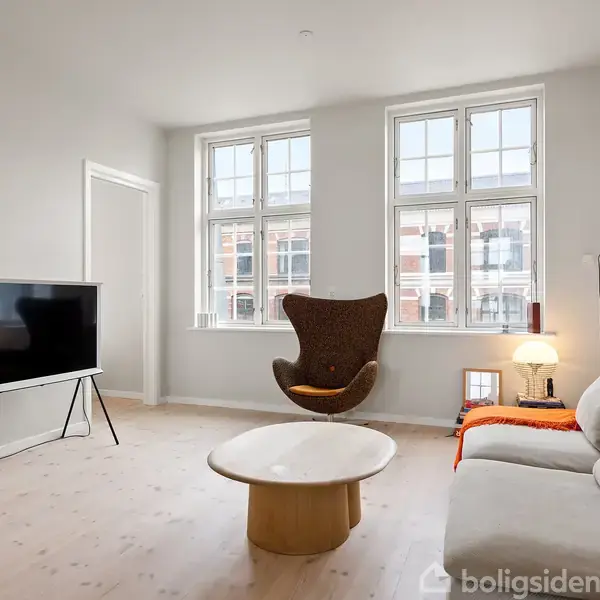 En lys stue med en sofa og bord som sidder stille ved siden af en væglampe, fjernsynet står tændt på et stativ, med store vinduer i baggrunden.