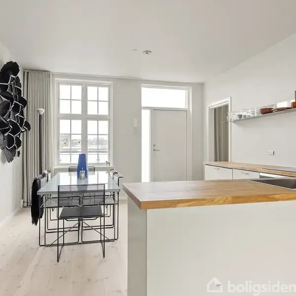 Køkkenbord med vask pryder en lys, minimalistisk stue med spisebord og stole. En skulpturel vægdekoration hænger til venstre, og store vinduer modtager naturligt lys i rummet.