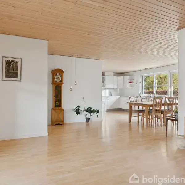 Urmøbel står i hjørnet og viser tiden; et spisebord med stole står nær store vinduer. Lyse trægulve og træloft skaber en lys, åben stue-alrum.