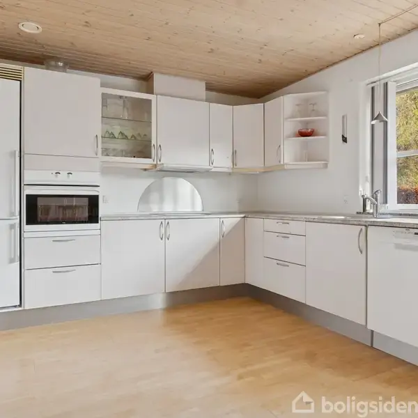 Et hvidt køkken med moderne skabe og indbygget ovn, står i et lyst rum med trægulve og et stort vindue, der giver udsigt til grøn bevoksning udenfor.