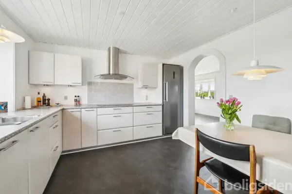 Et moderne køkken med hvide skabe, en marmorbordplade og en emhætte. Et spisebord med en vase blomster står til højre. En buegang fører til en tilstødende stue.