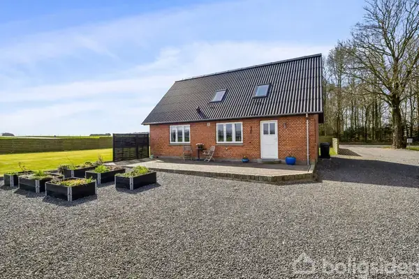 En rødstensvilla med sort tag står på en grusbelagt gårdsplads. Foran huset er der flere plantekasser og en grøn græsplæne. Træer ses i baggrunden til højre.