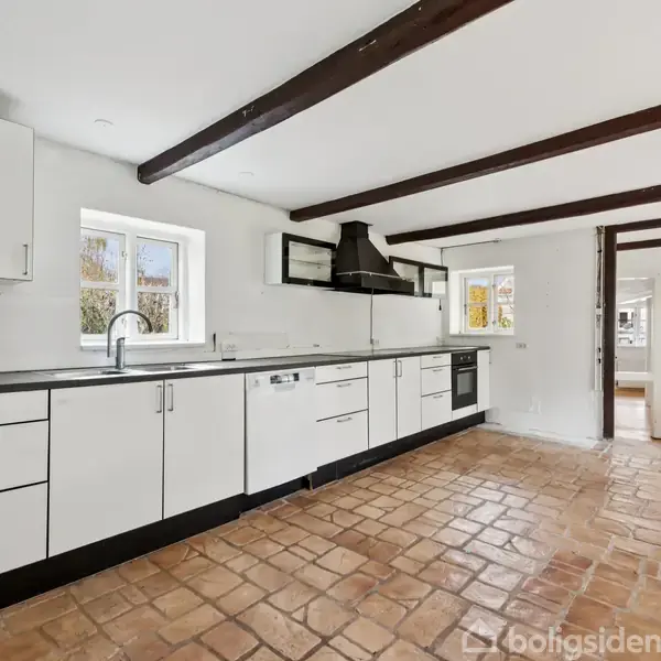 Et moderne køkken med hvide skabe og sort bordplade står langs en væg med to vinduer. Keramiske fliser dækker gulvet, og synlige bjælker pryder det lyse rum.