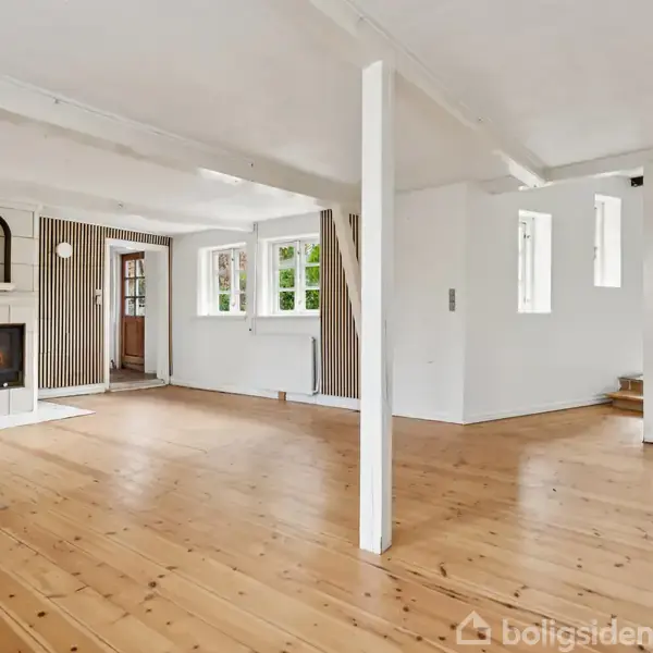 Stue med træbræddegulv, pejs med ild i, omgivet af hvide vægge og bjælker. Store vinduer lader lys ind og en trædør fører til den tilstødende del af huset.