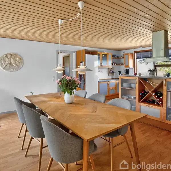 Et køkken med et træbord er omgivet af seks stole. En brændeovn står i hjørnet, og der hænger lamper over bordet. Et køkkenområde er bagerst med moderne apparater.
