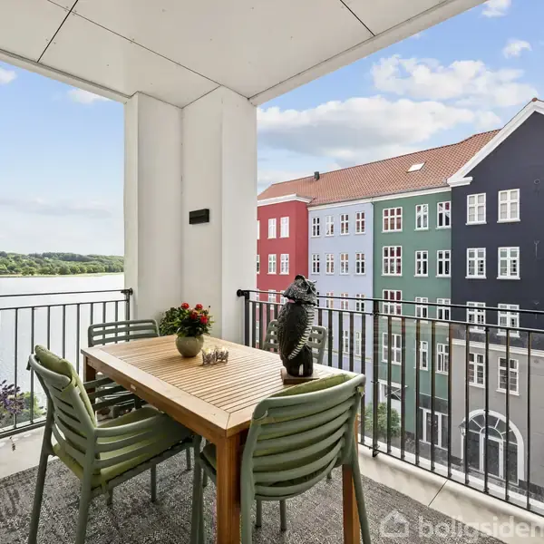 En balkon med et træbord og fire stole står stille, med udsigt til en malerisk række flerfarvede bygninger og en sø i baggrunden.