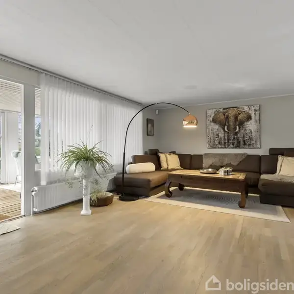 Brun hjørnesofa med puder står på en trælaminatgulv i hyggelig stue med kunstværk af elefant på væggen og gulvlampe. Glasdør fører ud til lys veranda.