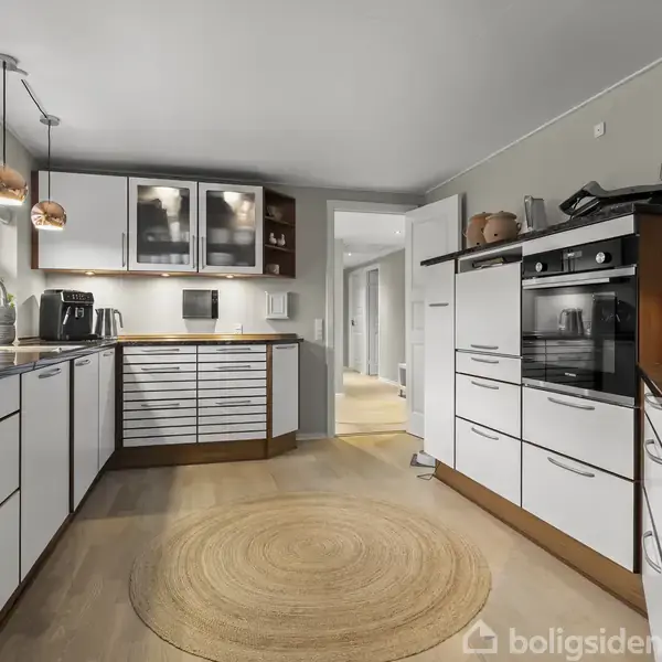 Et moderne køkken med hvide skabe og sorte bordplader, er udstyret med indbyggede apparater. Rund tæppe på trægulvet; vindue til venstre viser udendørs landskab.