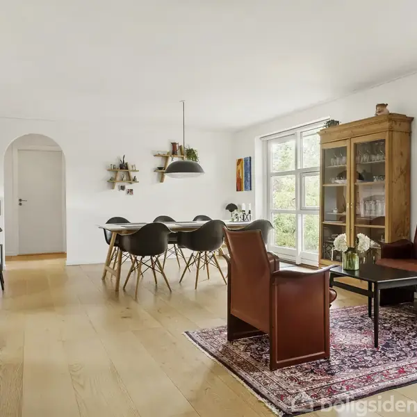 Stue med moderne møbler og trægulv, en brun lædersofa ved et sofabord på et orientalsk tæppe, spisebord med stole, vitrineskab og store vinduer, der giver naturligt lys.