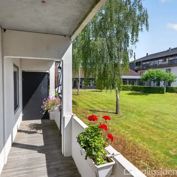 En altan med blomsterkasser står stille mod en grøn græsplæne. I baggrunden ses et hvidt byggeri og træer under en blå himmel med spredte skyer.