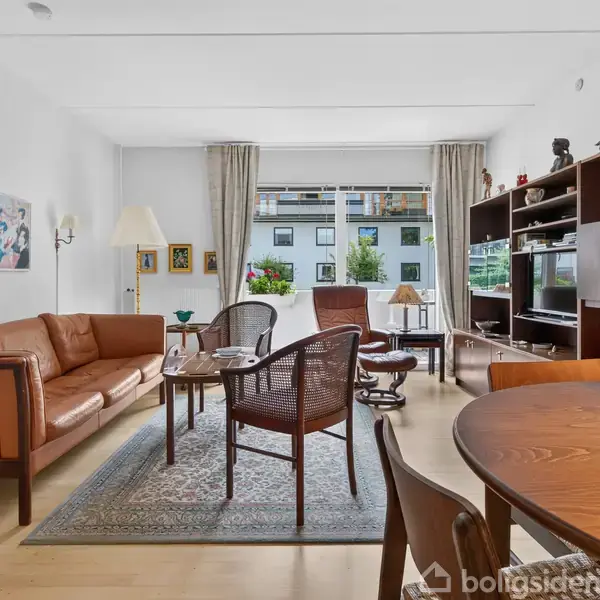 Stue med lædersofa og stole omkring et tæppebord, bogreol med tv. Store vinduer med udsigt til altan og grønne planter. Træmøbler og vægkunst tilføjer elegance.