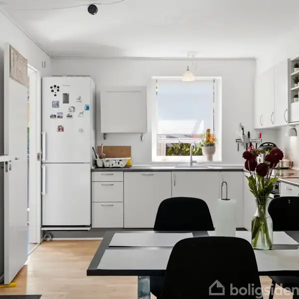 Køkken med hvide skabe og sorte stole omkring et spisebord; et køleskab med magneter står til venstre, og et vindue med en plante er bagud.