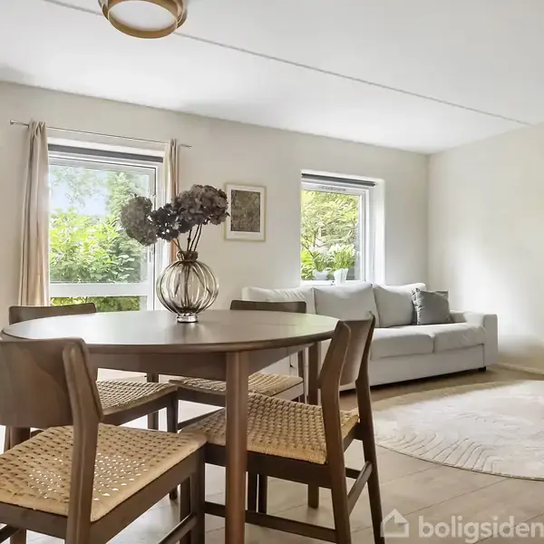 Et rundt træbord står centralt i en stue med fire stole omkring. I baggrunden ses en sofa, store vinduer med kig til grønne omgivelser, og en bogreol med planter.