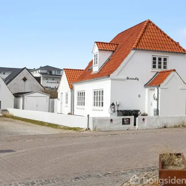 Hvidt hus med rødt tegltag står stille på en brostensbelagt vej. Nummer 8 og navnet "Strandly" er skrevet på væggen. Bagved ses flere huse med lignende arkitektur.