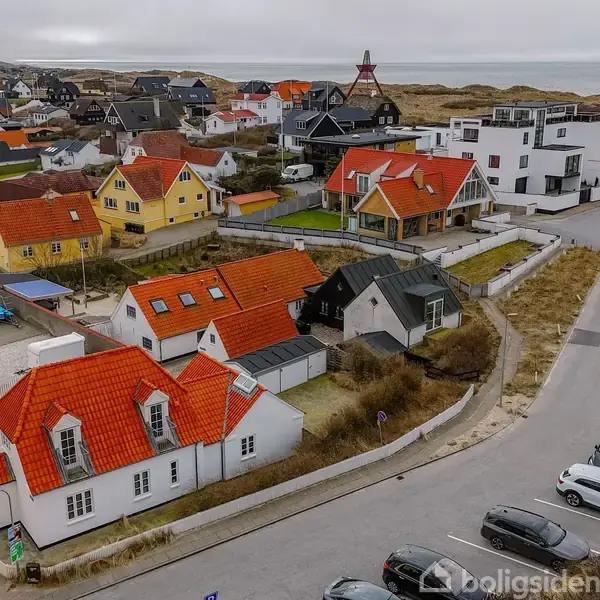 Huse med røde tage står stille i en kystby nær klitter og hav. Omkringliggende bygninger har forskellig farver, hvor flere biler er parkeret ved en vej.