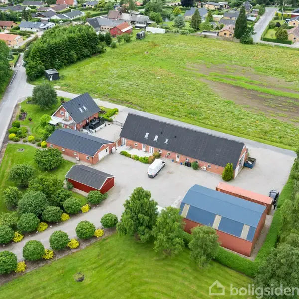 Et stort gårdanlæg med røde bygninger omgivet af grønne marker og træer. Et køretøj står parkeret på en gårdsplads. En vej løber langs siden med flere huse i baggrunden.