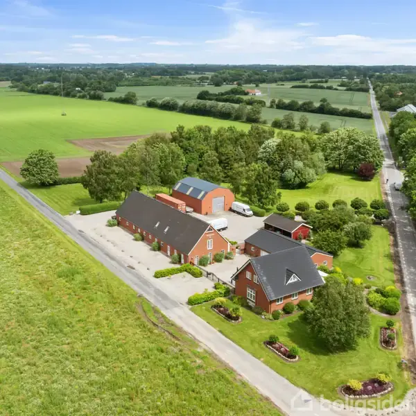 En række røde landejendomme står langs en vej omgivet af grønne marker og træer, placeret i et landligt område under en blå himmel med spredte skyer.