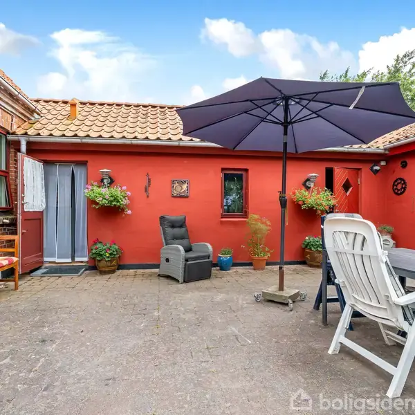 En gårdhave med et bord, stole og en stor parasol på en flisebelagt terrasse. Omkring er der røde vægge og skodder, samt blomsterkrukker og en bænk under en blå himmel.