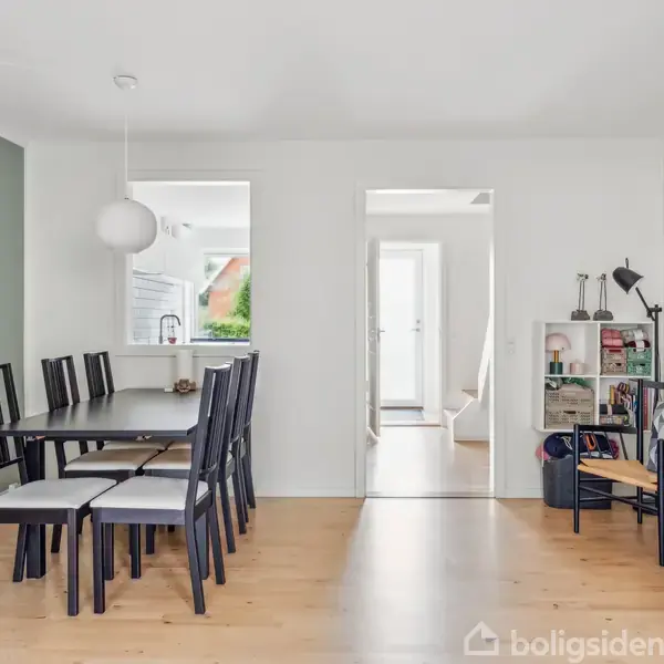 Spisebord med sorte stole står i et spisestueområde med lyst trægulv. Et hvidt, rundt loftlampe hænger over. I baggrunden ses en åbning til et andet rum med dagslys.