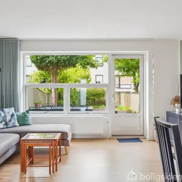 Grå sofa med puder står i en stue, hvor der er et TV på et møbel og et stort vindue med udsigt til en frodig have.