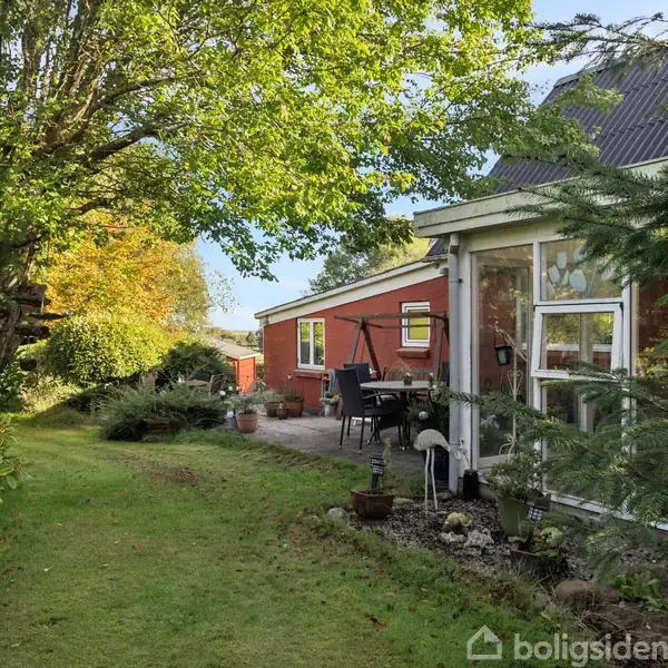 En rød murstensbygning med en terrasse møbleret med stole og planter. Træer og buske omkranser haven, der skaber en fredfyldt atmosfære.