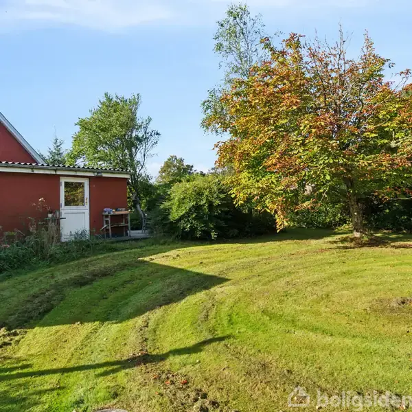 En rød bygning med hvide døre og vinduer står ved siden af en grøn have, hvor et stort træ kaster skygger på den velplejede græsplæne.
