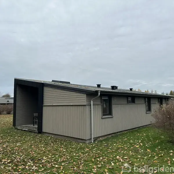 Grå træbygning står stille på en græsplæne dækket af spredte efterårsblade, omgivet af buske og træer under en overskyet himmel.