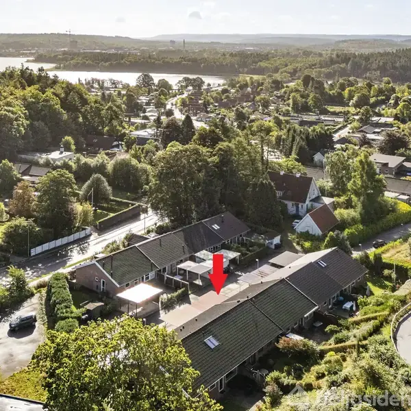 Rækkehus markeret med en rød pil set ovenfra, omgivet af træer og andre huse i et villakvarter ved en skov og sø i baggrunden.