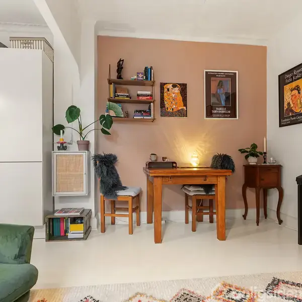 Et skrivebord med to stole står op ad en væg med kunstplakater. Et grønt sofahjørne med en farverig pude er til venstre. Reoler og plante ses også.