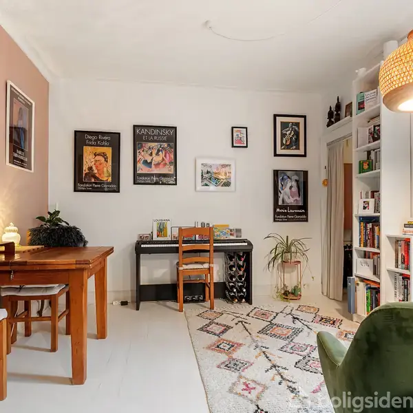 Et hyggeligt indrettet værelse med et træbord og stole ved en pink væg. Et klaver med en stol står op ad en hvid væg dekoreret med kunstplakater. En gylden lampe hænger over et mønstret tæppe. Reoler med bøger og planter fylder rummet.