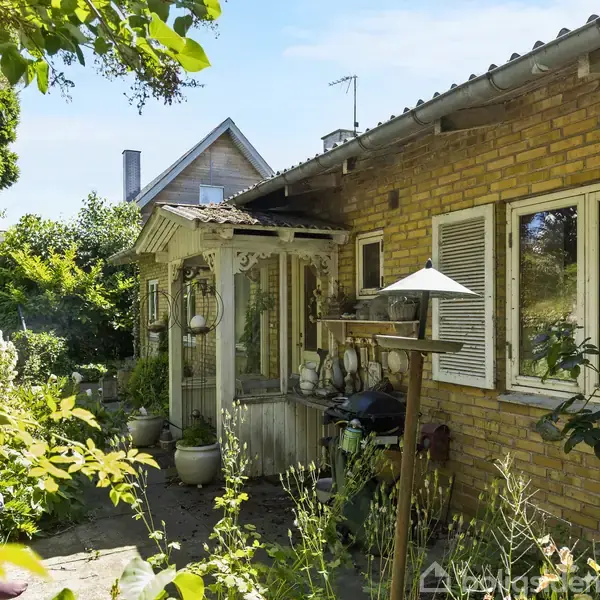 En gul murstensvilla har en terrasse fyldt med potteplanter og grønne buske i en frodig have. Et mindre trætag beskytter indgangen, og solen skinner over det hele.