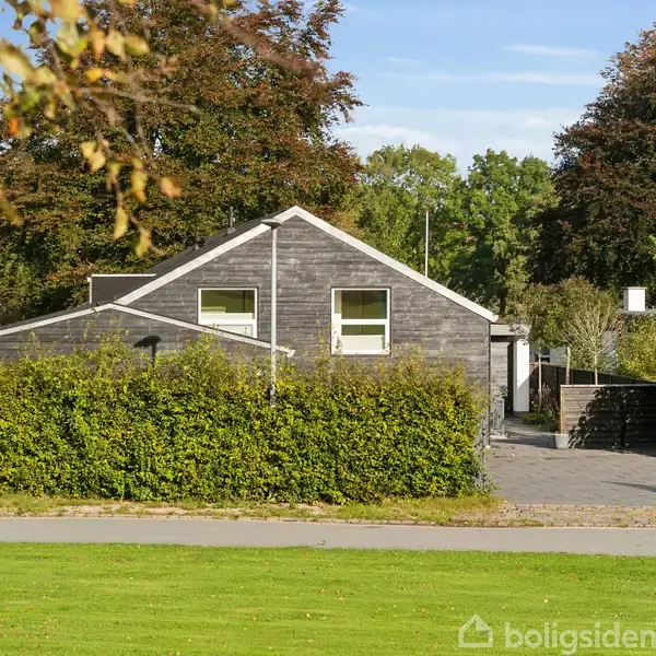 Huset står stille omgivet af en hæk og indkørsel, med klare vinduer mod den blå himmel. Store træer omkranser området i en fredelig forstad.