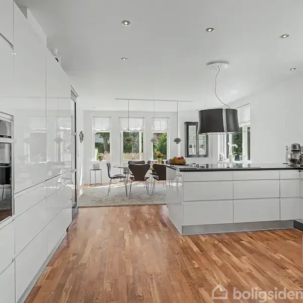 Køkken med hvide, moderne kabinetter og en sort emhætte, åbner til en lys spisestue med store vinduer og træbord. Trægulvet giver varme til det minimalistiske design.
