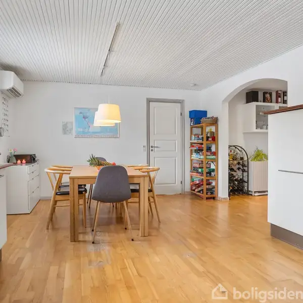 Spisebord med stole stående i et køkken-alrum. Bordet har en lampe hængende over sig. Rummet har trægulv, hvide skabe og døren fører til et tilstødende rum.